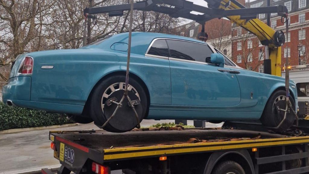 A blue Rolls Royce hangs suspended in the air as it is loaded on to a relocation vehicle. 