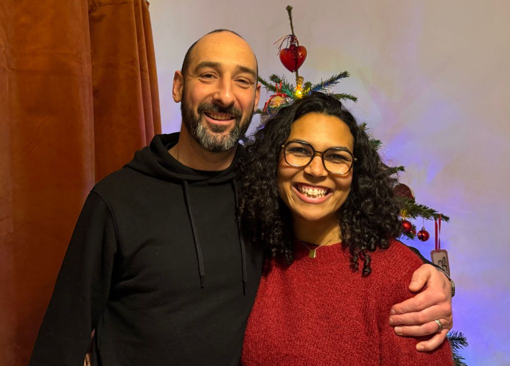 Daniel in black hoodie with his arm around Rachel in red jumper an glasses. Both are smiling and a Christmas tree is behind them