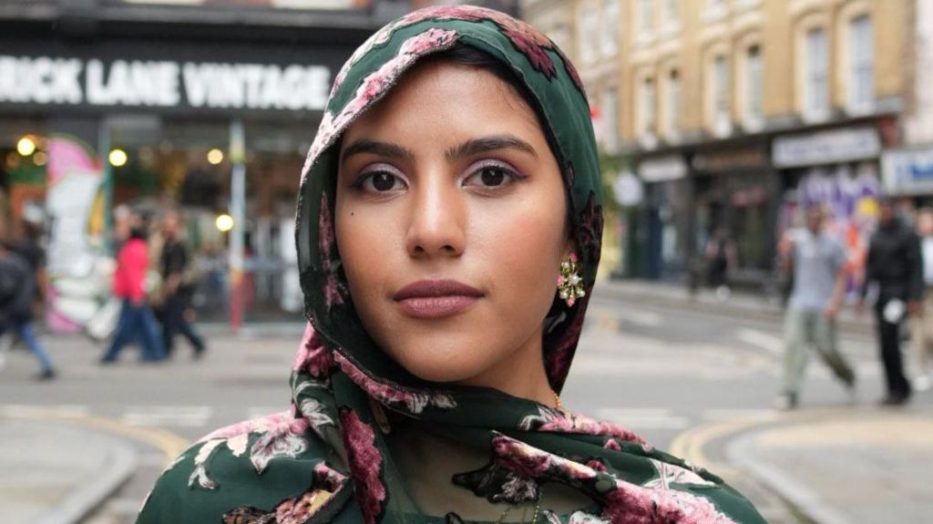 Nabeela Zaman wearing a green and pink floral hijab, in the background is a high street area, blurred out slightly.