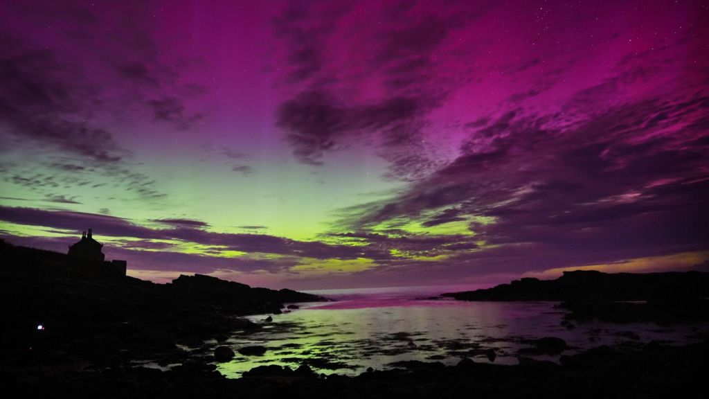 Aurora borealis in Howick, Northumberland, showing the sky in layers of purple, indigo and various shades of green, with hilly terrain and river below in silhouette