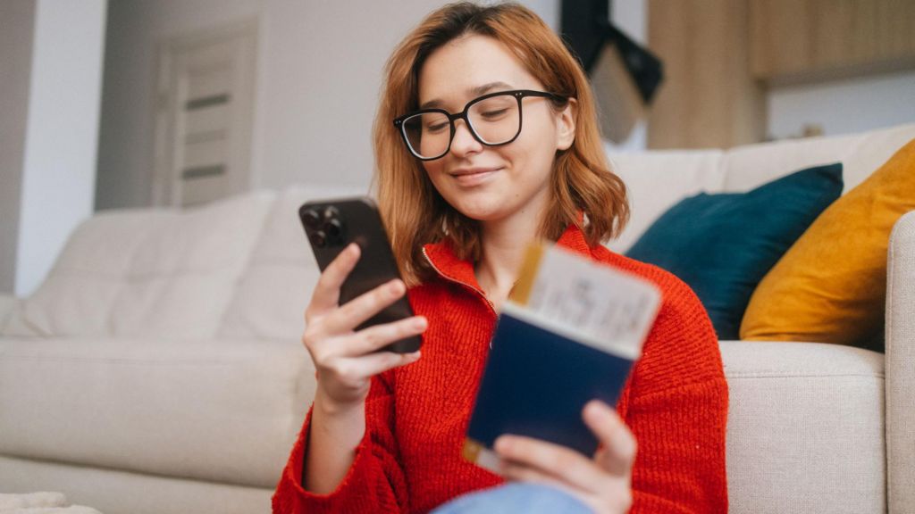 A young woman looks at her phone. She is holding her passport and a plane ticket in her other hand.