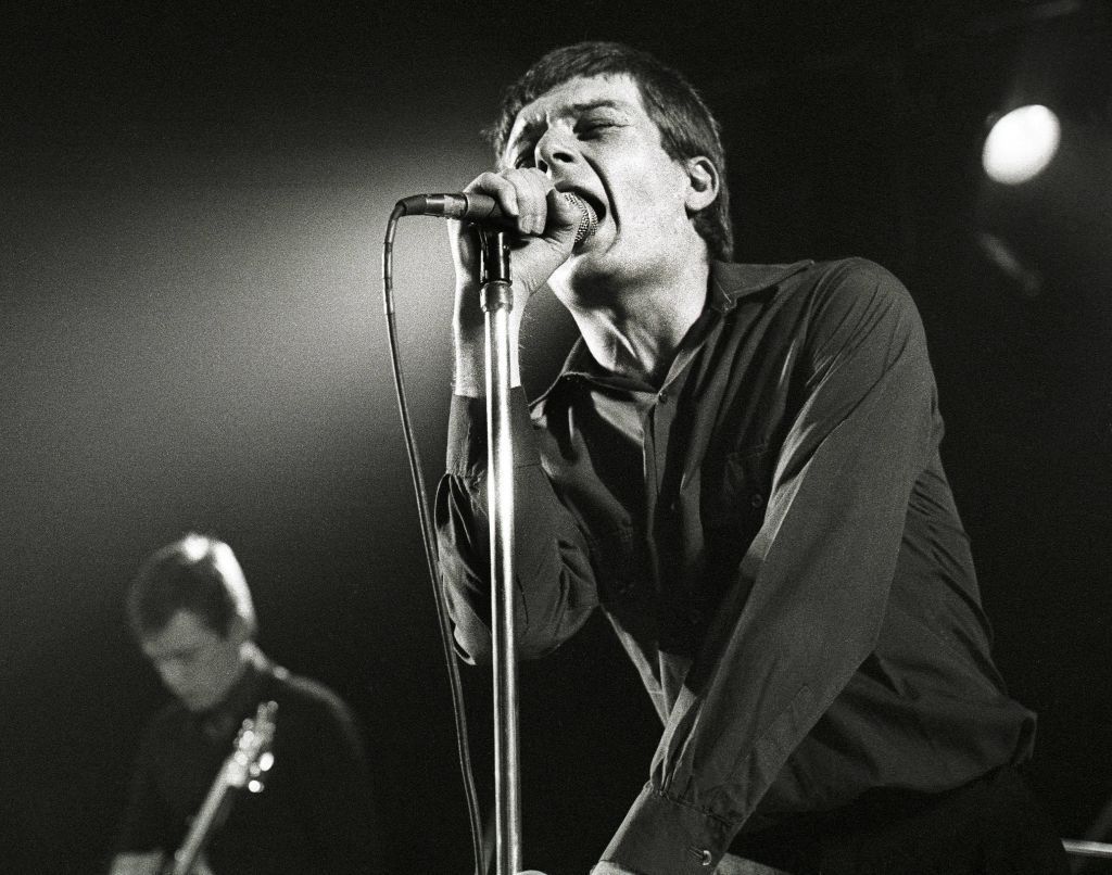 Ian Curtis of Joy Division, pictured in 1980, singing into the microphone on stage with his eyes closed.