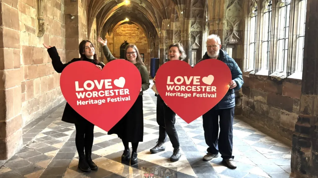 Three women and one man in a corridor with a vaulted stone roof and stone-framed windows holding two large heart-shaped signs which read 