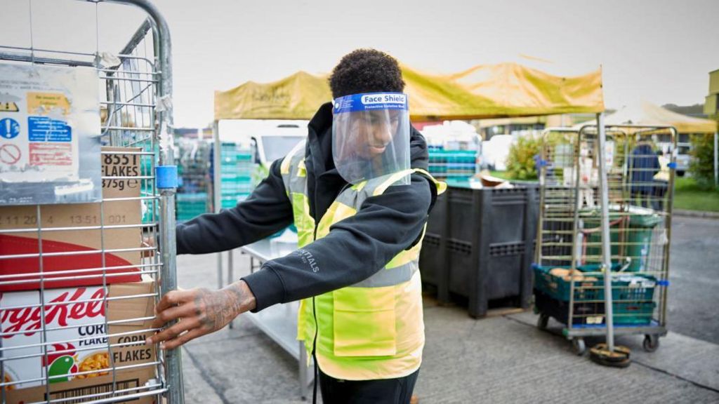 Marcus Rashford in a high-vis vest, wearing a face shield, pulls a warehouse cage full of Cornflakes 