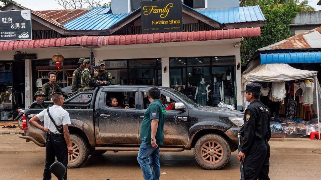 A military vehicle inside the city. Armed men in fatigues stand on the back of a black pickup truck driving down a road with shops on it, one called Family Fashion Shop