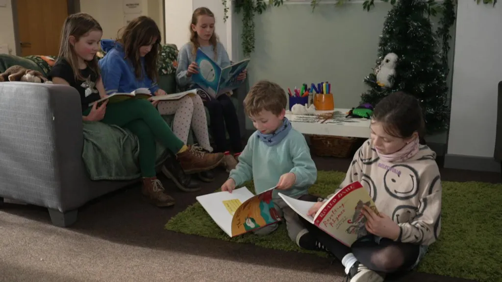 The picture shows a cosy reading area where several children are sitting together with books. Some are settled on a sofa while others are on the floor on a green rug, each absorbed in their own story. The space is decorated with a small tree and greenery along the wall, as well as a low table with colourful pens and craft materials. 