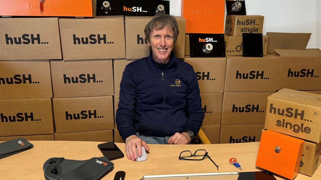 Paul Nugent sitting at a wooden desk with piles of cardboard boxes printed with Hush on them behind him.