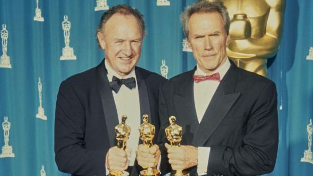 Gene Hackman and Clint Eastwood, both in tuxedos, with Hackman holding an Oscar in his right hand, and Eastwood holding an Oscar in each hand. They are standing in front of a blue curtain and a life-sized version of the Oscar statue can be partially seen in the background.