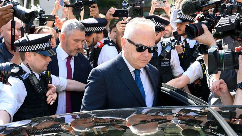 Huw Edwards looks down at the floor while wearing black sunglasses, as he gets into a black car surrounded by police and paparazzi on a sunny day outside Westminster Magistrates' Court on 31 July