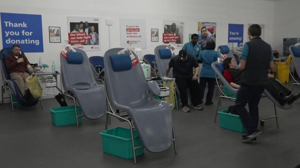 The blood-giving room in a hospital, with donors sitting in big chairs as they give blood. A number of the chairs are empty and the wall bears posters thanking people for donating.
