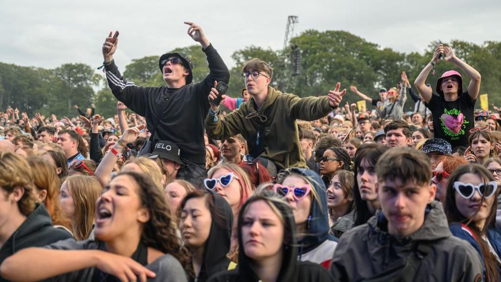 A crowd of people stand in daylight watching someone perform at Leeds Festival. Three men sit on people's shoulders watching the music.