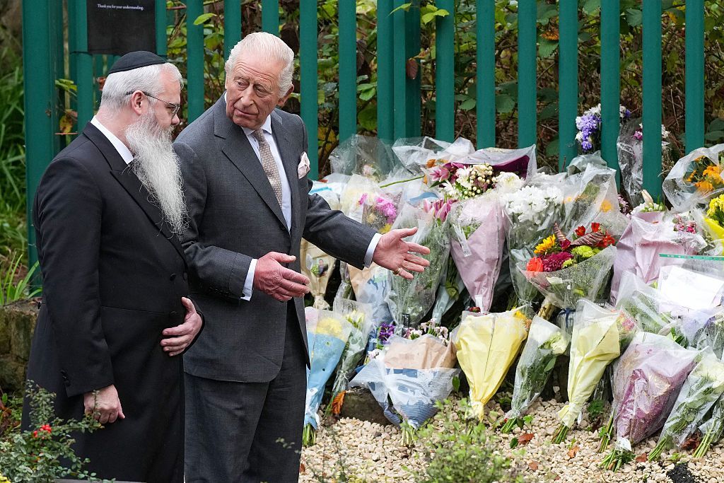 A man with a long white beard, white hair and glasses is with another man (King Charles III) - both are wearing suits. King Charles's hands are outstretched towards more than a dozen bunches of flowers laid on the ground against a fence.