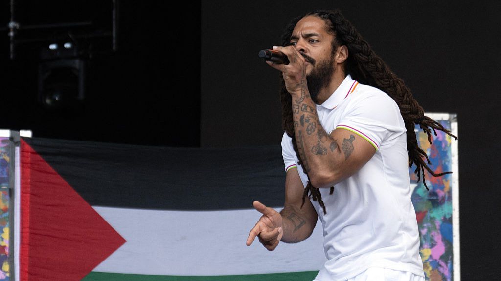 Bobby Vylan, a man with long dreadlocks, performs with a microphone while standing in front of a Palestine flag.