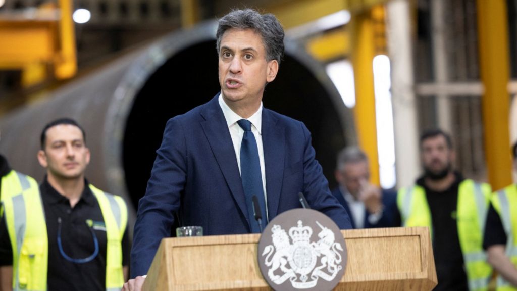 Energy secretary Ed Miliband stands behind a lectern with a government crest and speaks to workers at an industrial site 