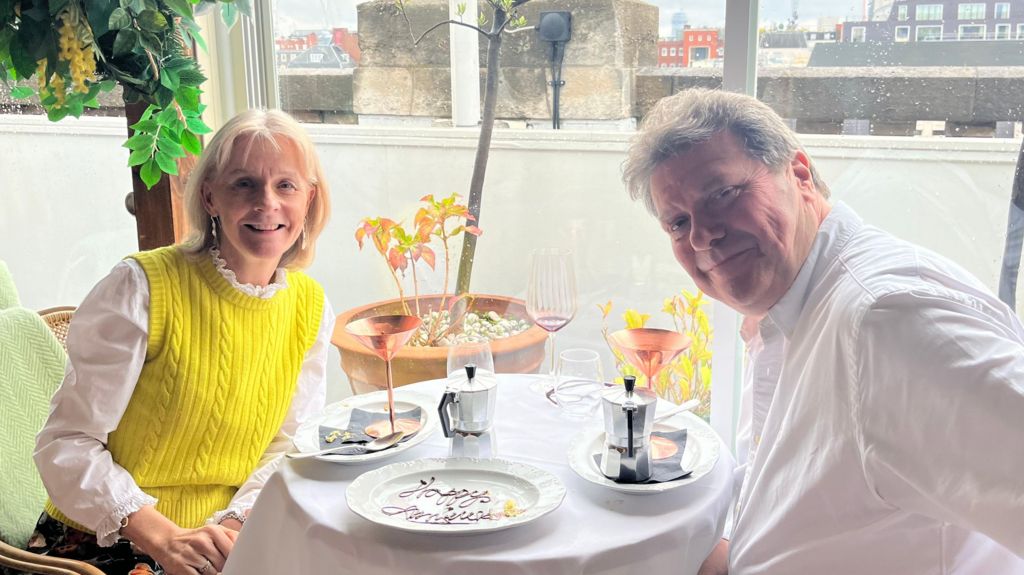 Sue and Alex sit at a table in front of a window, a plate saying 'Happy anniversary' in chocolate between them. They smile at the camera, Alex's arm outstretched Sue is wearing a yellow sleeveless jumper over a white shirt and earrings, she has blonde hair. Alex is wearing a white shirt and his hair as turned grey. There are two pots of what looks like coffee at the table and small plants and buildings in the background.