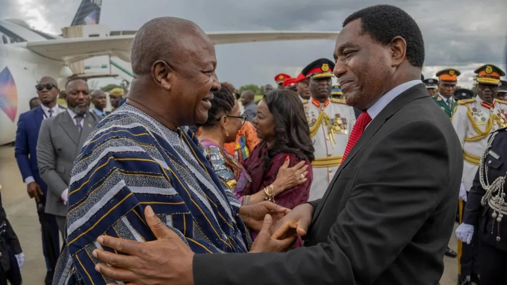Zambia President Hakainde Hichilema greets Ghana President John Dramani Mahama warmly on an airport tarmac.  Mahama is wearing a traditional striped Ghanaian fugu and Hichilema is in a dark suit with a red tie. They stand in the middle of a welcoming procession, that includes uniformed military personnel, dignitaries, and an aircraft in the background..