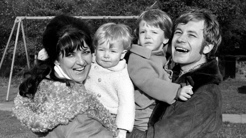 A woman holds one child and a man holds another in a black and white image. The four smile and laugh and there is a playground in the background.