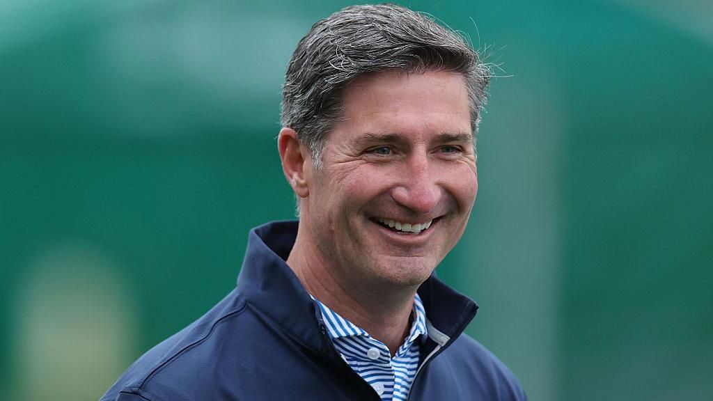 Starbucks chief executive Brian Niccol, smiling and wearing a striped blue and white shirt under a dark blue jacket with the collar up.