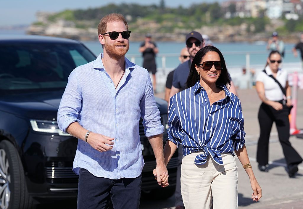 Harry and Meghan walk together hand in hand in front of a beach in Australia.