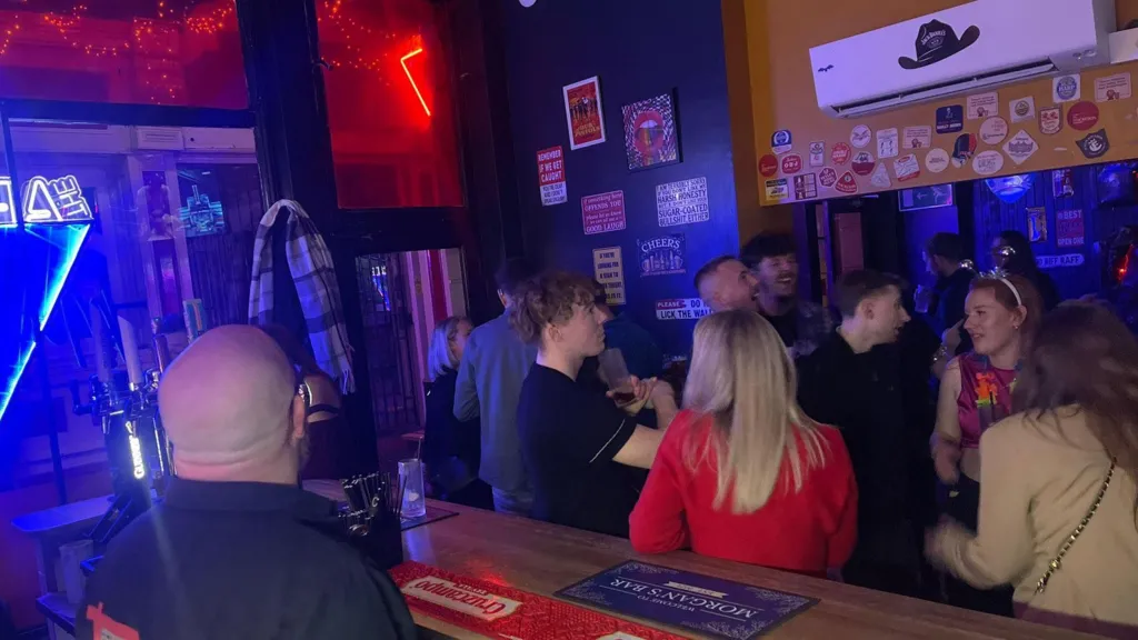People gathered at Morgan's bar where they are drinking and mingling. Morgan is in a black shirt standing behind the bar, which has branded bar mats and drink taps. The walls are decorated with signs, stickers and beer mats, and blue and red neon lighting illuminates the room.
