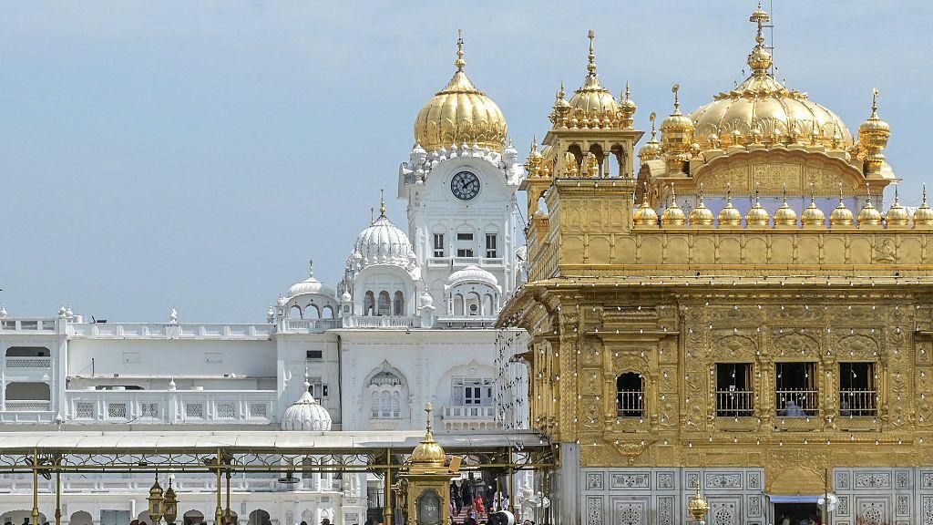 A view of the Golden Temple complex in Amritsar, Punjab. The gold-covered Harmandir Sahib dominates the foreground, while white marble buildings with domes and a clock tower rise behind it under a clear blue sky. The temple is the holiest shrine in Sikhism and is visited by millions of pilgrims each year.