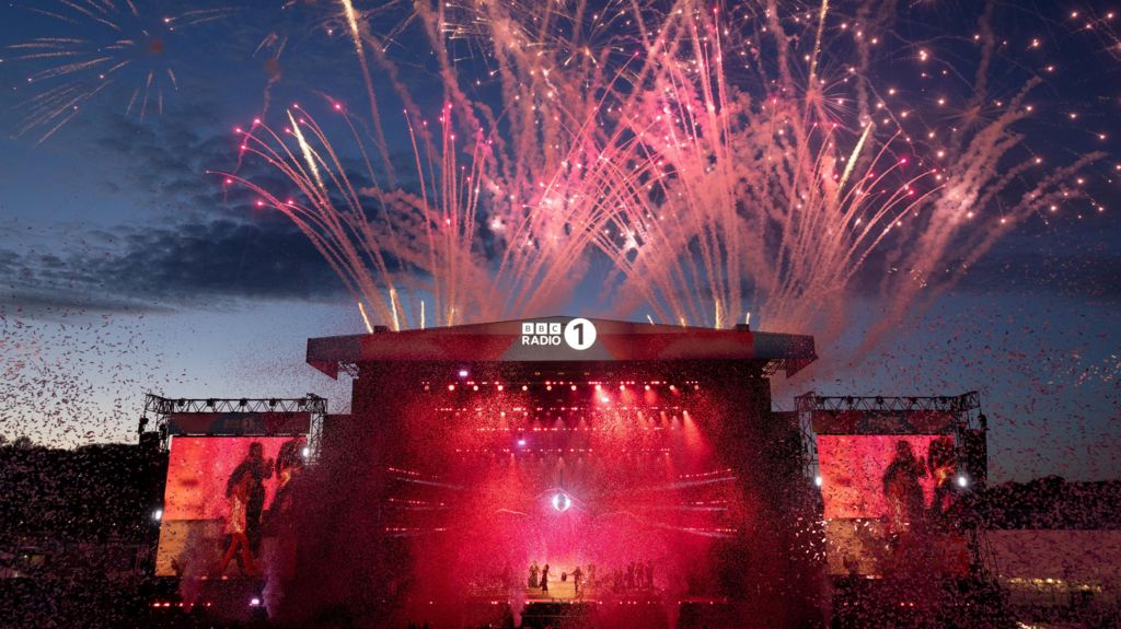 Olivia Dean is seen on stage on a screen, with pink lights and smoke in the sunset.