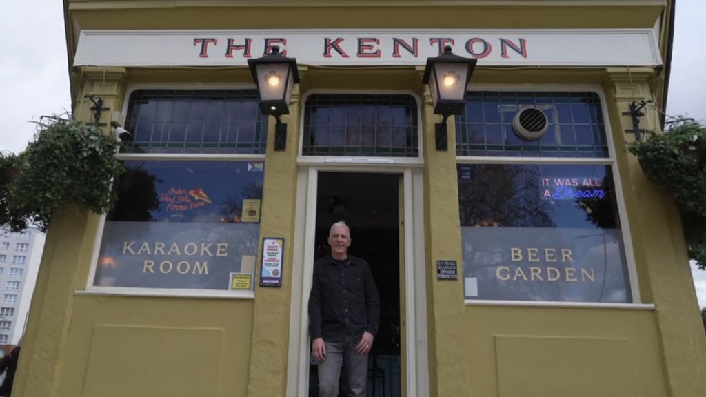 The pub is a yellowish/green colour with old fashioned signage that reads The Kenton and lamp lights. Signage in the windows reads "karaoke room" and "beer garden". A man with grey hair and wearing a black shirt and grey jeans stands in the doorway. The camera is pointing at an angle upwards from the ground.