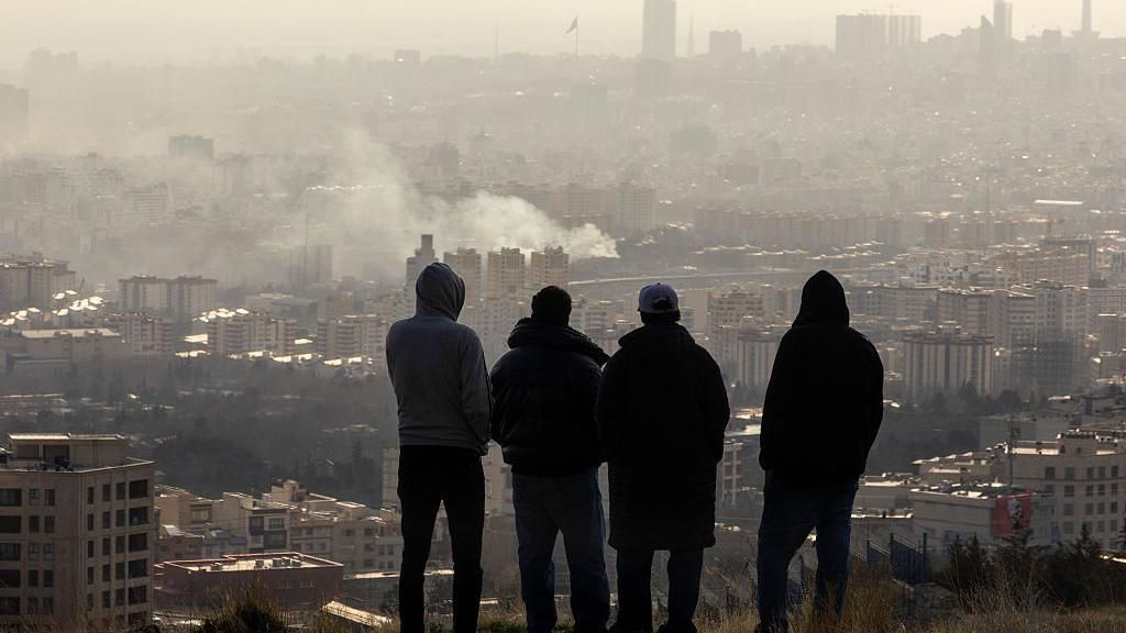 Men watch from a hillside as smoke rises after an explosion in Tehran, Iran, March 2026