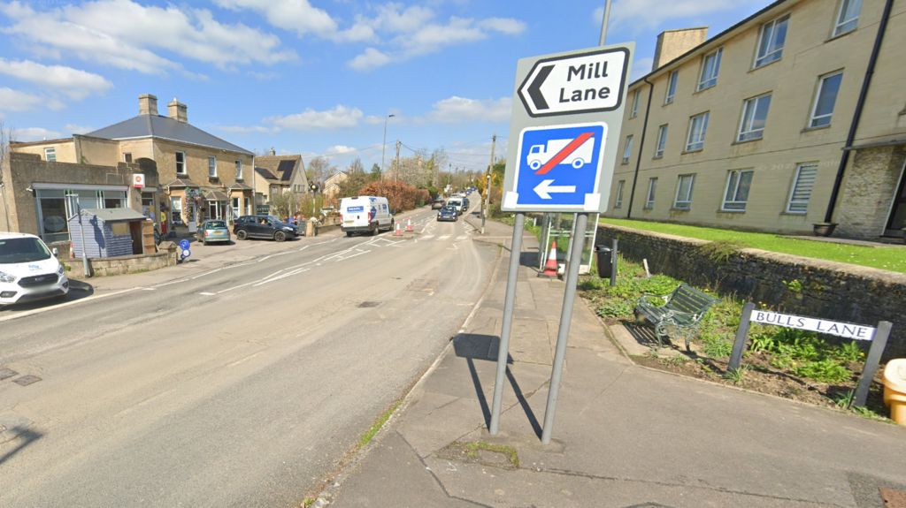 A signboard with two signs, one indicating Mill Lane and the other that there is no access for lorries. The signboard is next to a row of houses on the right, a Post Office outlet on the left and a zebra crossing in the distance.