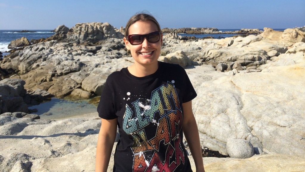 Tarryn Baird standing in front of rocks on a bench in front of the sea wearing a black T-shirt that says "glam slam".