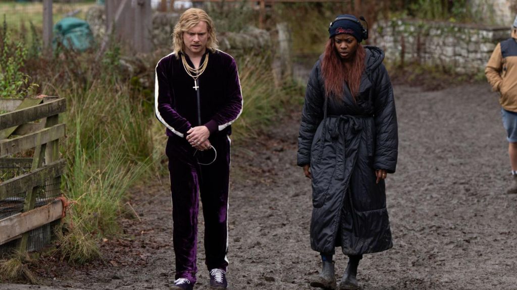 Jack O'Connell and Nia DaCosta on the set of 28 Years Later: The Bone Temple - he wears a purple velour tracksuit and gold chains and has shoulder length blond hair, she has a long black padded coat and short boots - they are looking down and walking outside on a muddy path 