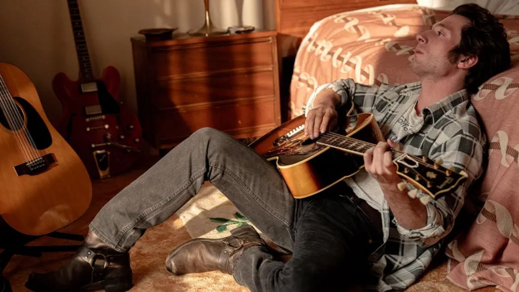 Jeremy Allen White as Brice Springsteen, sitting by a bed and playing an acoustic guitar