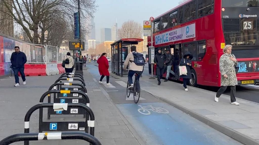 A floating bus stop in central London, with a cycle lane (marked in blue paint) runs between the pavement and a 'floating' area of pavement, on which the bus stop is located. There is a bus stop shelter and sign, at which a red double-decker bus is parked. A cyclist in the cycle lane is cycling towards a woman in a red coat, who is using a zebra crossing to cross the cycle lane to the bus stop.