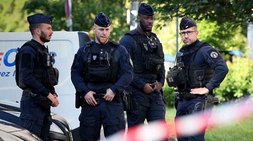 Four uniformed French police stand in front of a row of cars, with a police tape in the foreground, near the scene of the shooting of three boys in the city of Nantes.