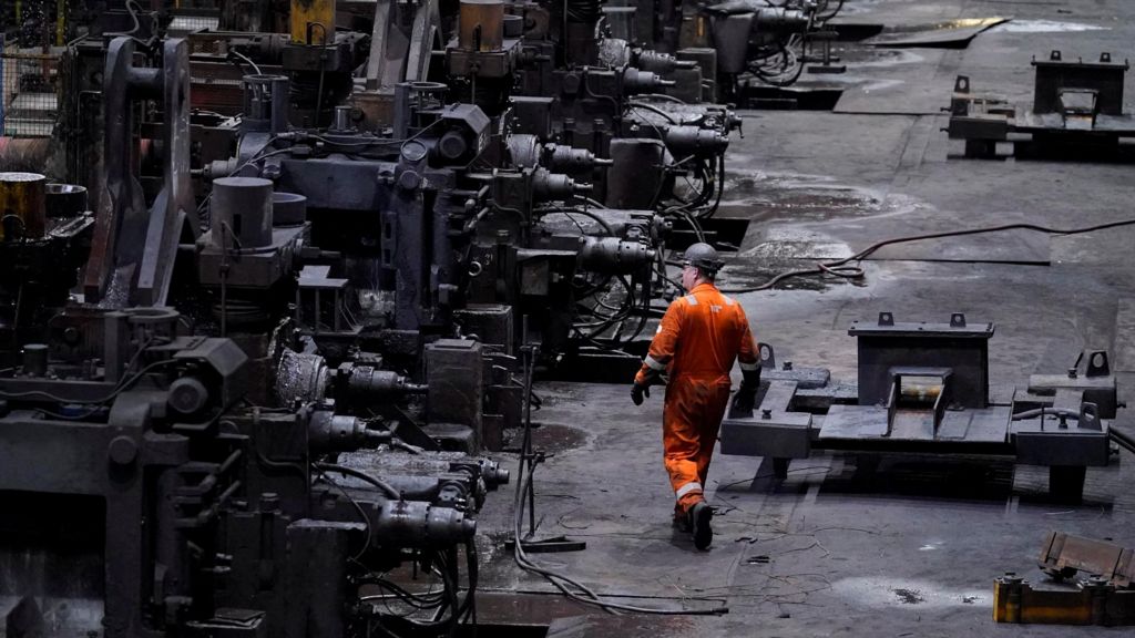 A wide view inside an industrial steelworks, showing long rows of large, dark metal machinery and workstations. A worker in bright orange protective clothing walks along the factory floor between the machines, with cables, metal parts, and heavy equipment scattered across the ground under dim industrial lighting.