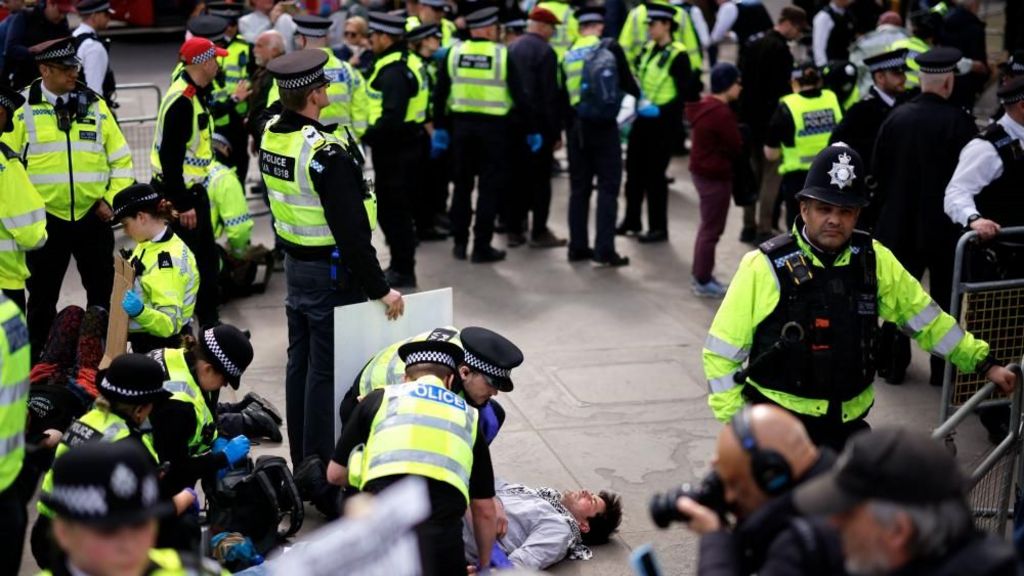A large group of police officers form a circle around a man in a grey sweatshirt lying on the floor. 