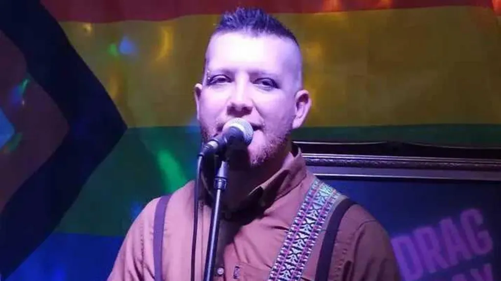 Patrick appears to be singing into a microphone, with blue lighting behind him. He has short spikey hair and a piercing in one eyebrow. He wears a brown shirt. He has a guitar strap over his shoulder.