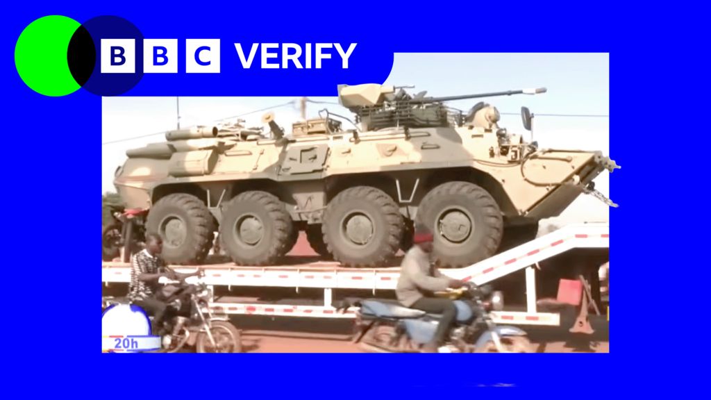 A Russian military vehicle being transported through the Malian capaitl, Bamako. Men on motorcycles are riding alongside it.