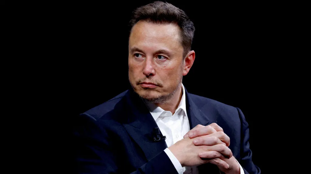 Elon Musk, empresario y propietario de X (anteriormente conocido como Twitter), con una expresión seria y las manos entrelazadas. Lleva un traje oscuro y una camisa blanca, y está sentado en un entorno con un fondo negro que resalta su figura.