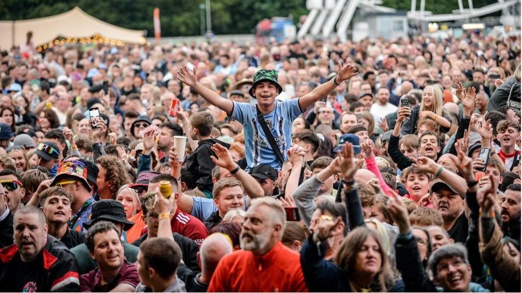 Godiva Festival crowd. We are focused on one man in the centre in a large crowd of people as he is sat on someone's shoulders while wearing a Coventry City FC shirt, a green hair and has a big smile with his arms outstretched towards the camera