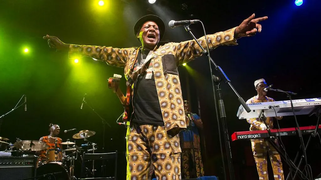 Ebo Taylor in a yellow African-print suit on stage. He is singing and has his arms stretched out either side of him.