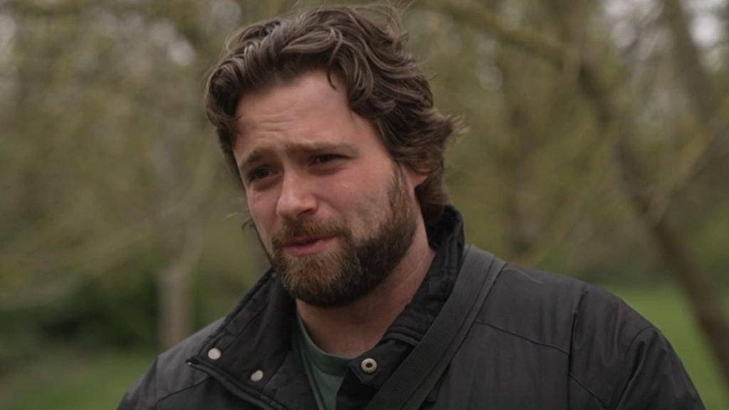 Kris Sikora is seen wearing a dark jacket stands outdoors in a park, with blurred trees in the background.