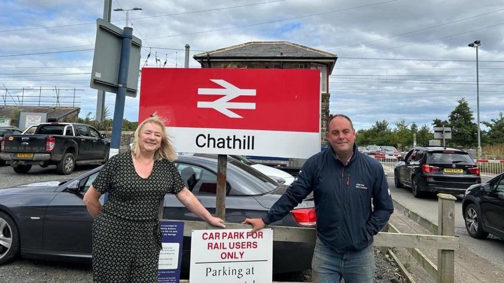 A photograph of two people standing in front of a big sign which says Chathill train station with the carpark and level crossing behind. 