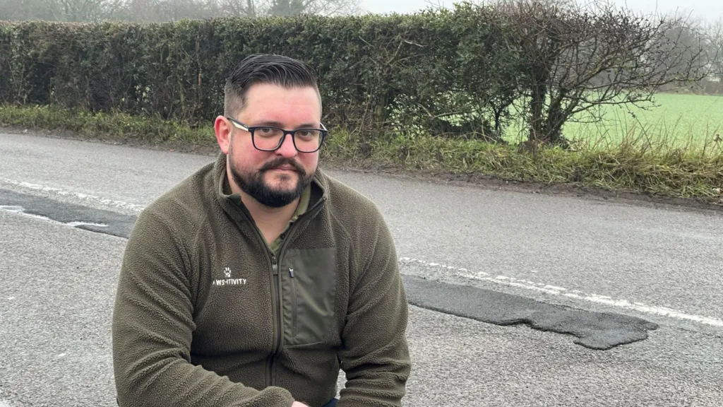 A man crouching on a pavement with a repaired pothole on the road in the background