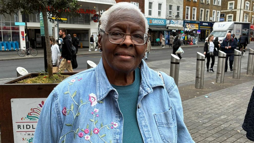 Valerie standing on a street wearing a blue denim jacket featuring a floral design. Shops like Greggs in the background.