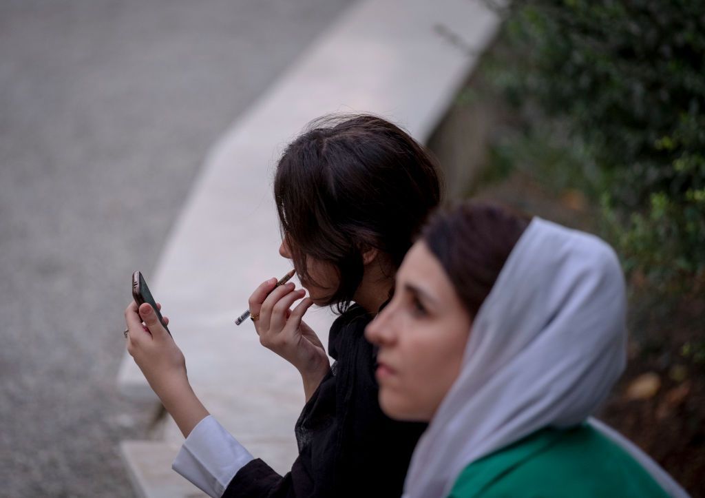 Two women sitting at a park bench. One is wearing a headscarf, the other isn't.