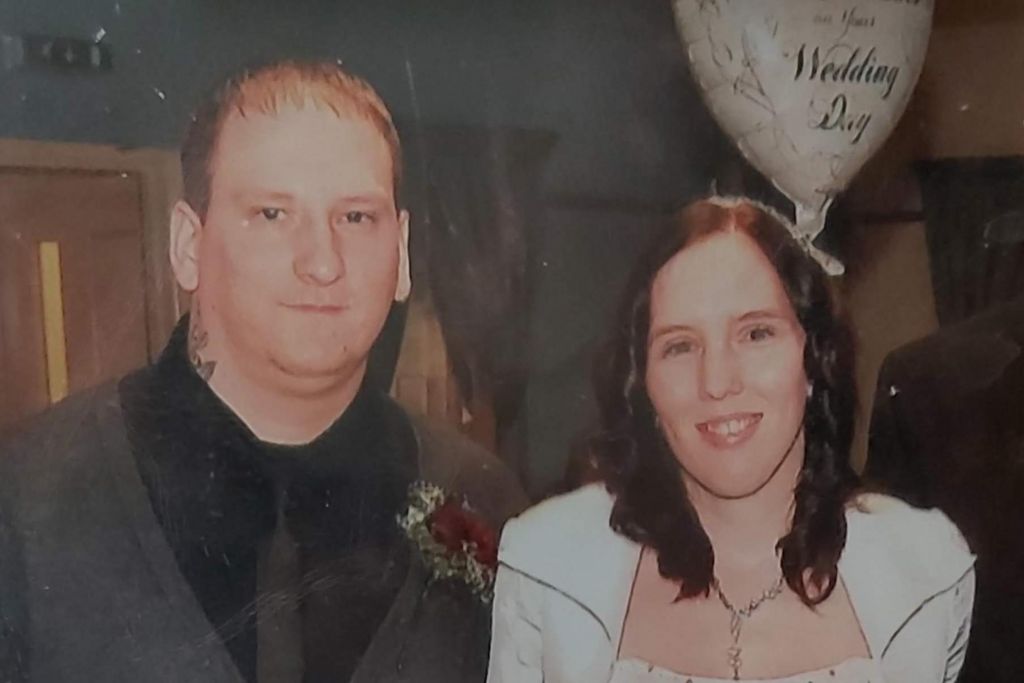 A man stands to the left of a woman. She is beneath a balloon which says "Wedding Day" and is wearing white. Her red-brown hair falls below her shoulders on to her front, where it touches a chain she is wearing. The man has thinning mousey hair which is cut short. He has a tattoo on his neck. He is wearing a black shirt and a dark grey tie and cardigan