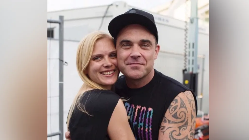 A woman with blonde hair is hugging Robbie Williams, who has his arms around her. They both smile at the camera.
