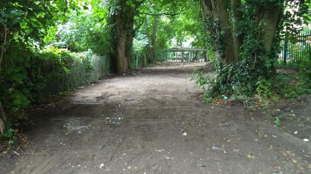 The site near The Brambles after being cleared. It is in a woodland area. There is a dirt track and lots of trees in the frame. 