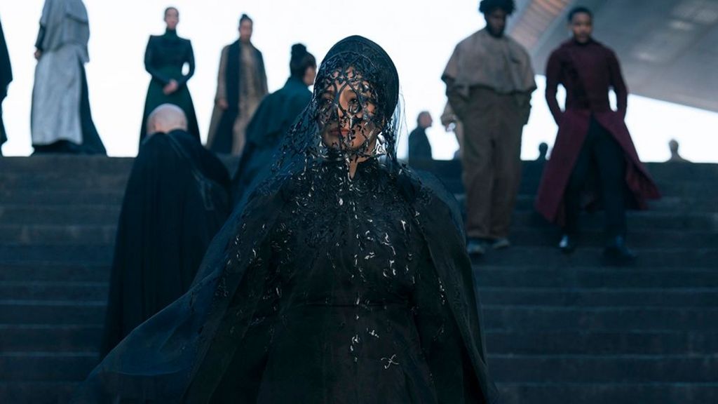 Tabu, playing the character of Francesca, dressed in all black, with a netted face veil, staring into the distance, while standing on dark coloured steps with people walking in the background.